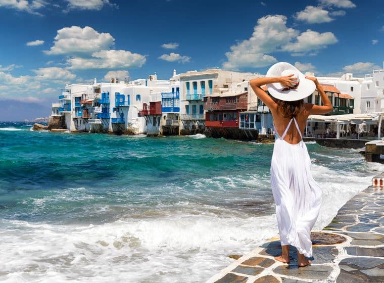 A woman in a white dress and hat looks out at the sea in Mykonos, Greece. White buildings with blue accents line the shore.