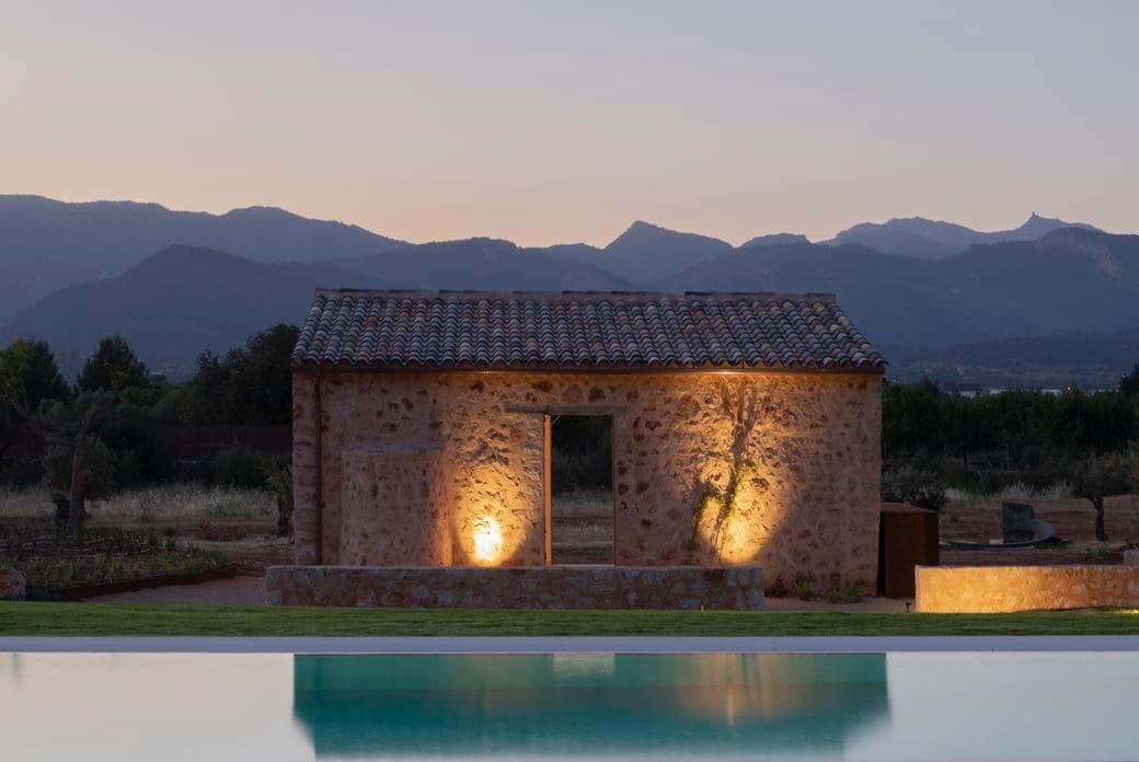 Un edifici rústic de pedra il·luminat al capvespre, amb una cadena muntanyosa al fons i una piscina reflectant al primer pla, situat a Santa Maria, Mallorca.