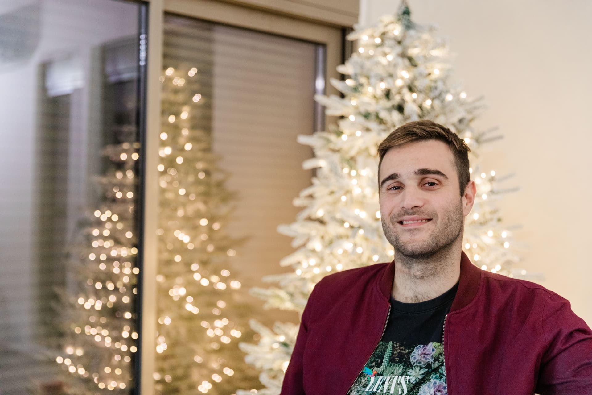 A smiling man in a maroon shirt stands in front of a white Christmas tree with lights.