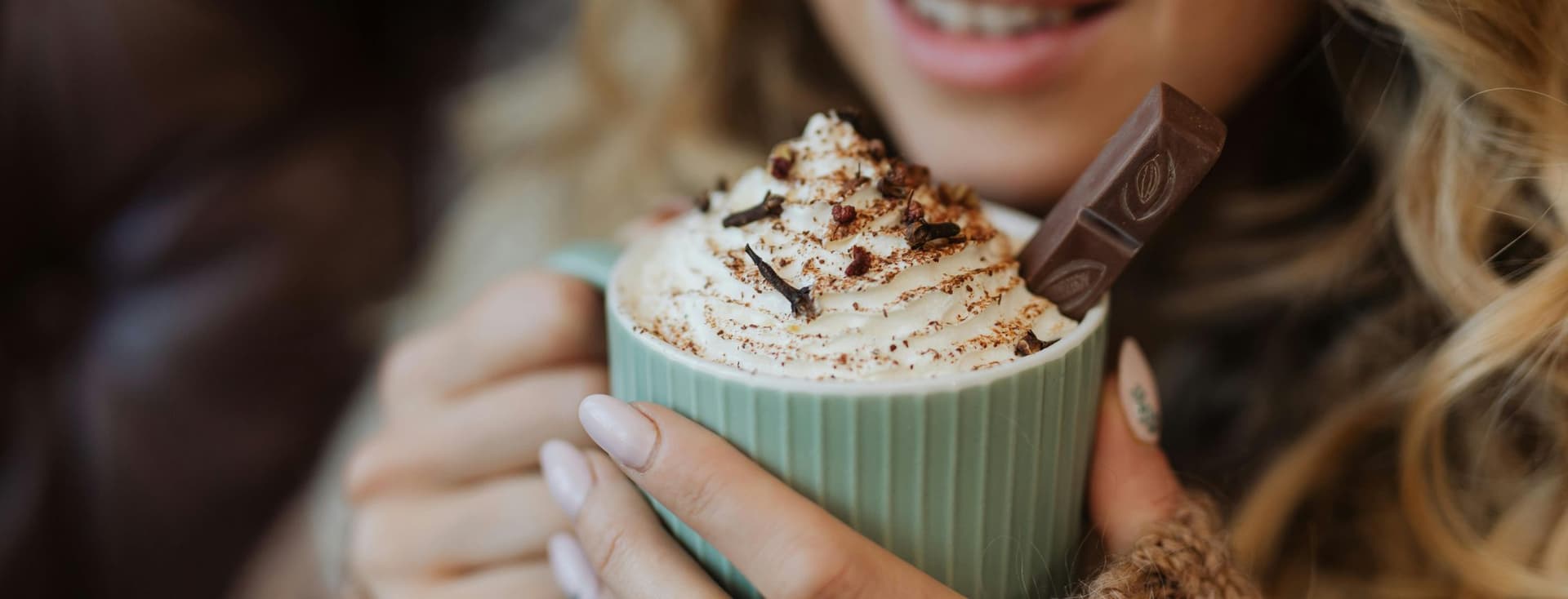 Une femme souriante tient une tasse verte de chocolat chaud avec de la crème fouettée et une barre de chocolat. Elle porte un pull marron.