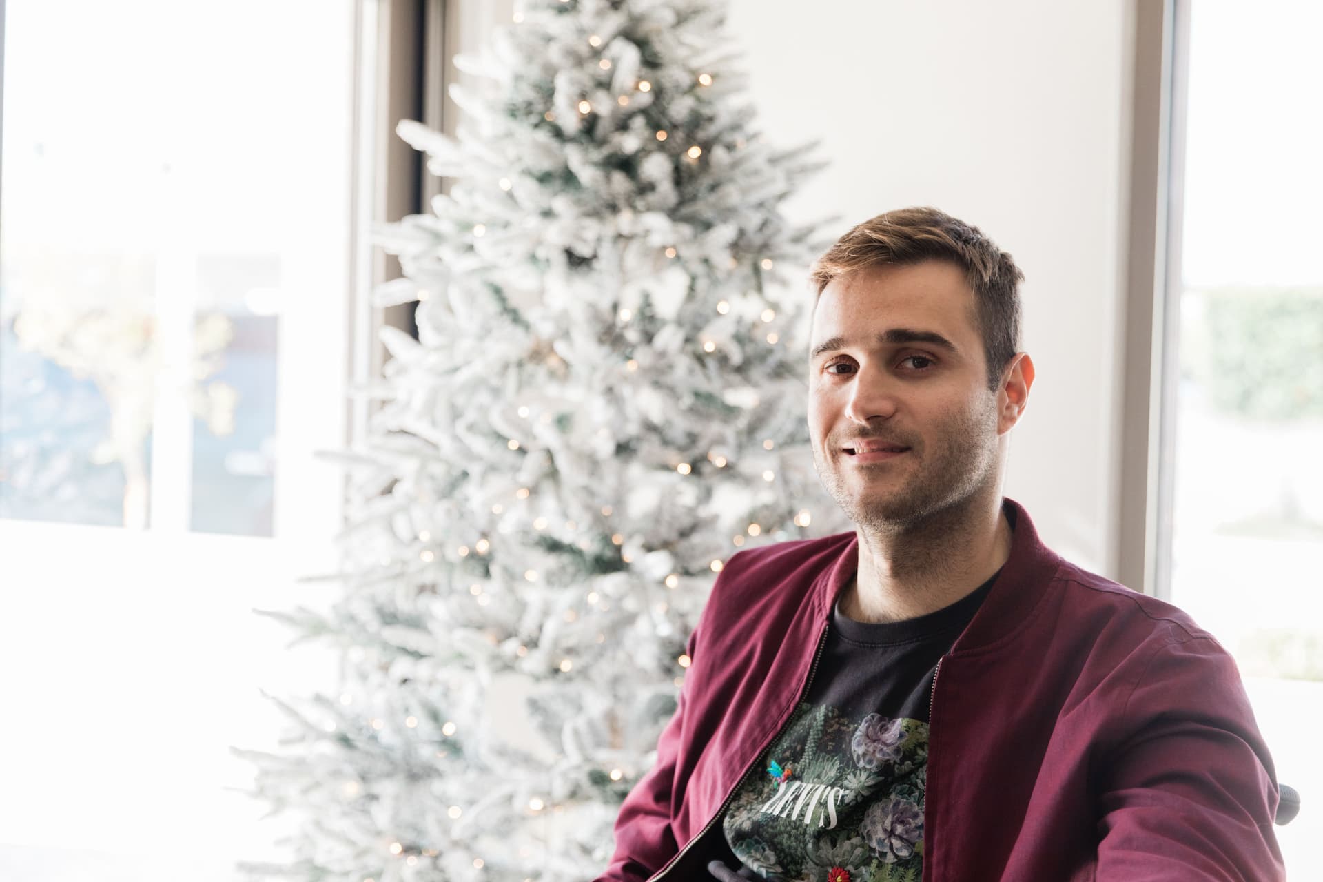 Man in a maroon jacket stands in front of a decorated Christmas tree.