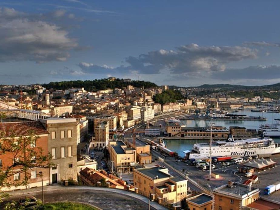Scenic view of a coastal city with historic buildings, a busy harbor, and a ferry docked under a partly cloudy sky.