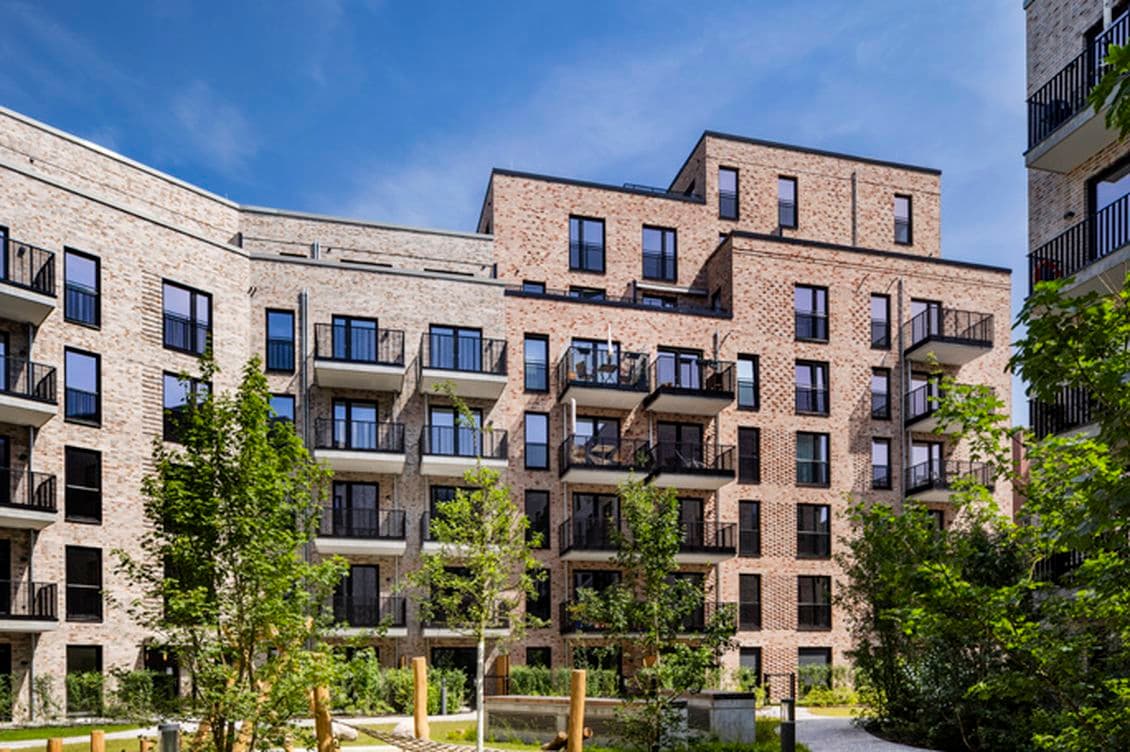 Modern brick apartment building with multiple balconies, surrounded by green trees and a small garden under a clear blue sky.