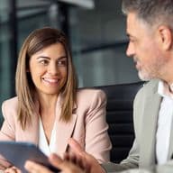 A smiling woman in a pink blazer holds a tablet while talking to a man in a gray suit.
