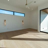 Bright, modern empty room with light wood flooring, large glass sliding doors to an outdoor patio, a ceiling fan, and a high horizontal window.