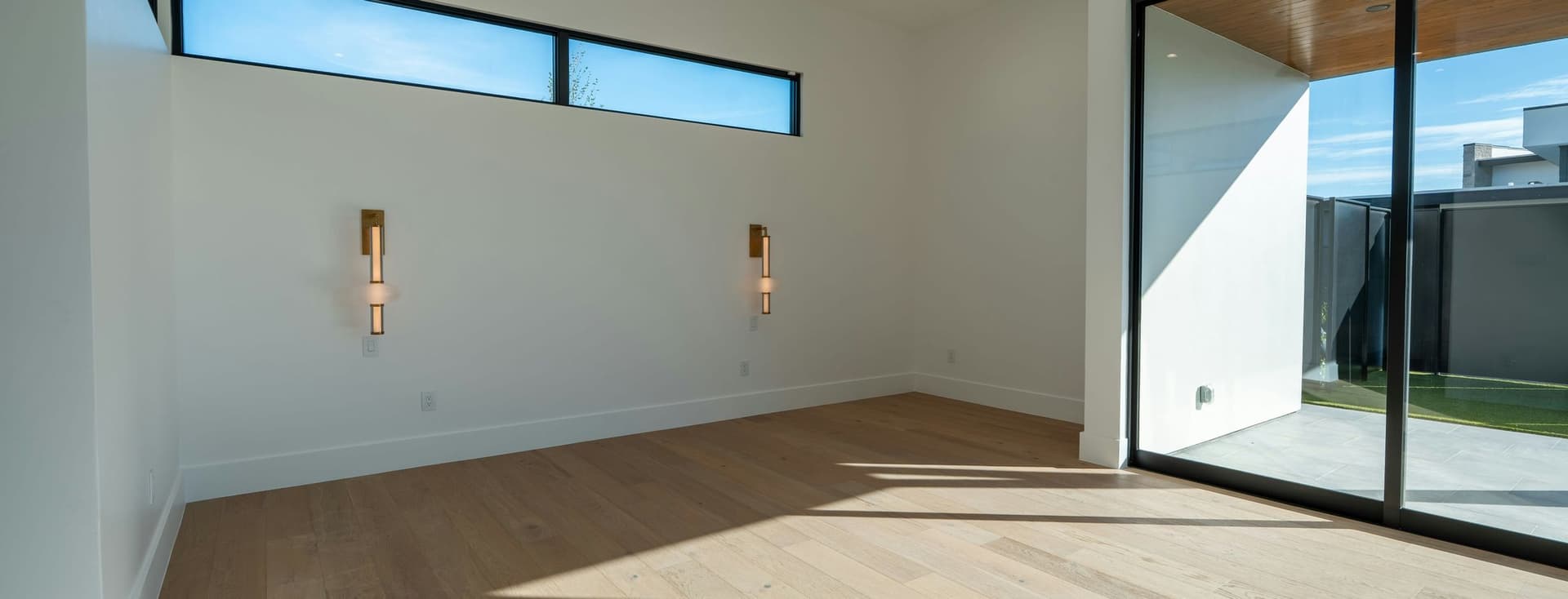Bright, modern empty room with light wood flooring, large glass sliding doors to an outdoor patio, a ceiling fan, and a high horizontal window.