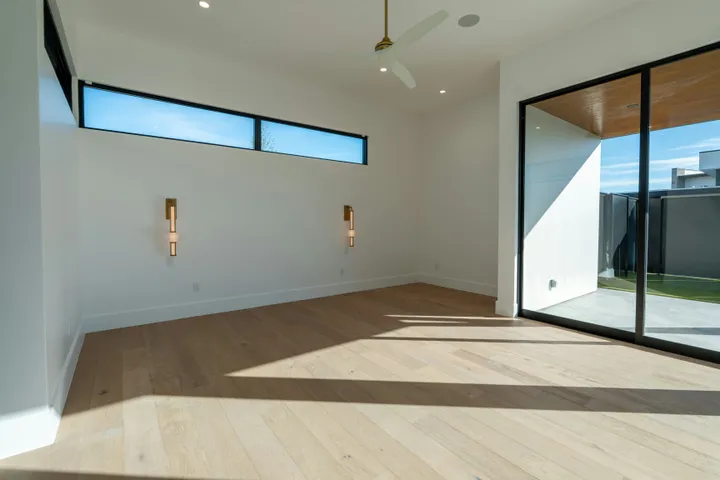Bright, modern empty room with light wood flooring, large glass sliding doors to an outdoor patio, a ceiling fan, and a high horizontal window.
