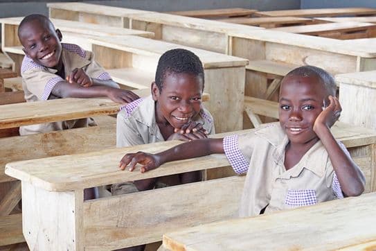 Three smiling children in uniform sit at wooden desks in a classroom, with two leaning on their desks and the third resting his head on his hand.