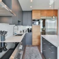 Sleek kitchen with gray cabinets, wooden pantry accents, stainless steel appliances, a white island, and two glass pendant lights.