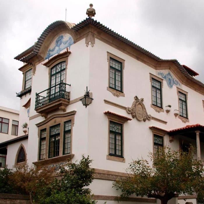 Ein elegantes zweistöckiges Gebäude mit dekorativen blauen Akzenten, Bogenfenstern und einem kleinen Balkon, umgeben von Bäumen unter einem bewölkten Himmel.