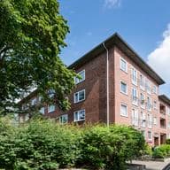 Red brick building with multiple windows, surrounded by green shrubs and trees under a blue sky with clouds.