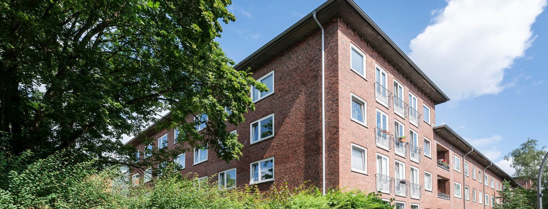 Red brick building with multiple windows, surrounded by green shrubs and trees under a blue sky with clouds.