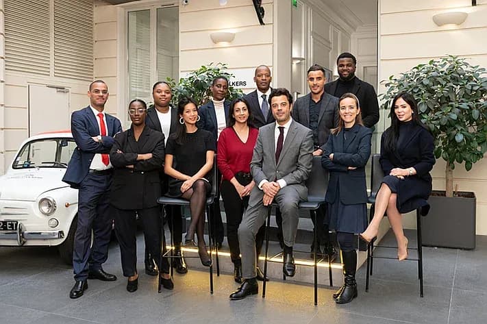 A real estate team of eleven poses in suits and dresses in front of a white vintage car.