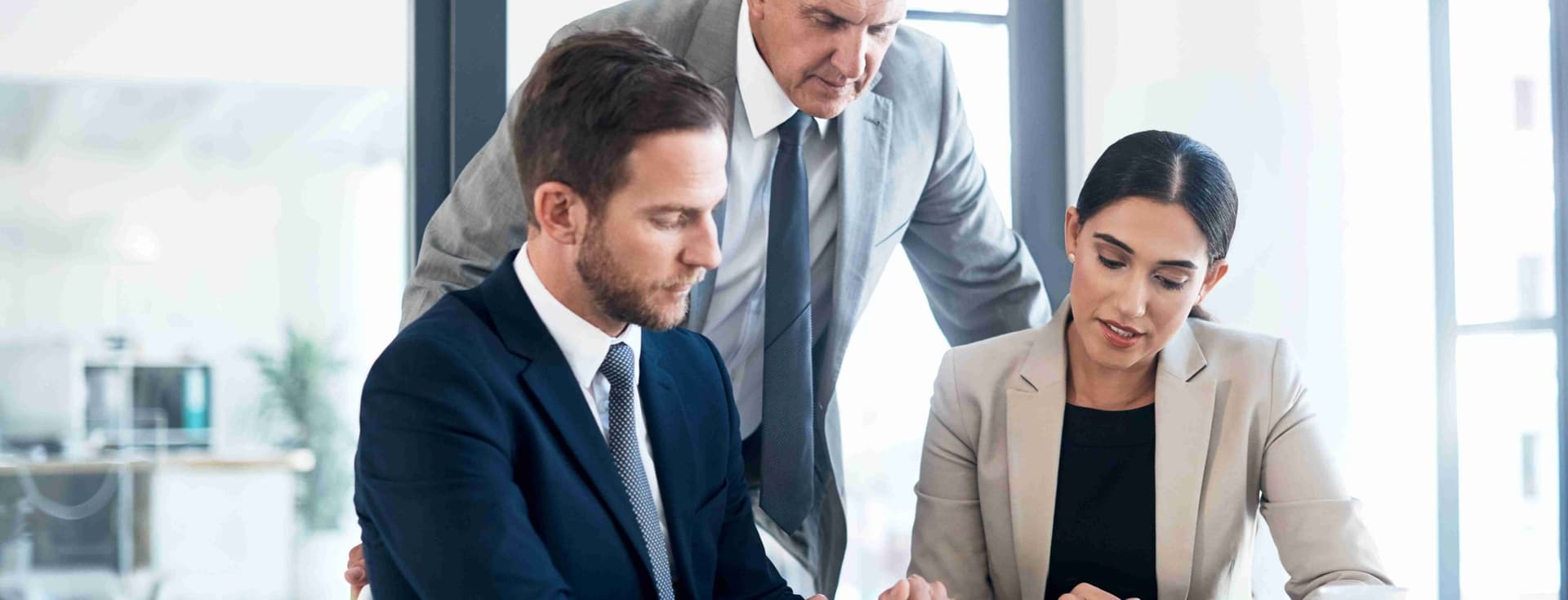 Three business people in suits review data on a tablet at a table in a bright office.