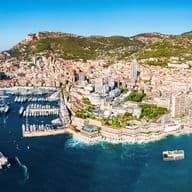 Aerial view of Monaco, a city-state on the French Riviera, with a harbor full of yachts and buildings stretching up a green hillside.