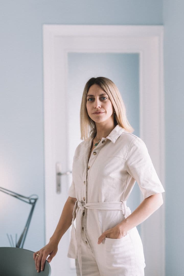 Μια ξανθιά γυναίκα με μπεζ φόρμα εργασίας στέκεται σε ένα γαλάζιο δωμάτιο με μια άσπρη πόρτα.