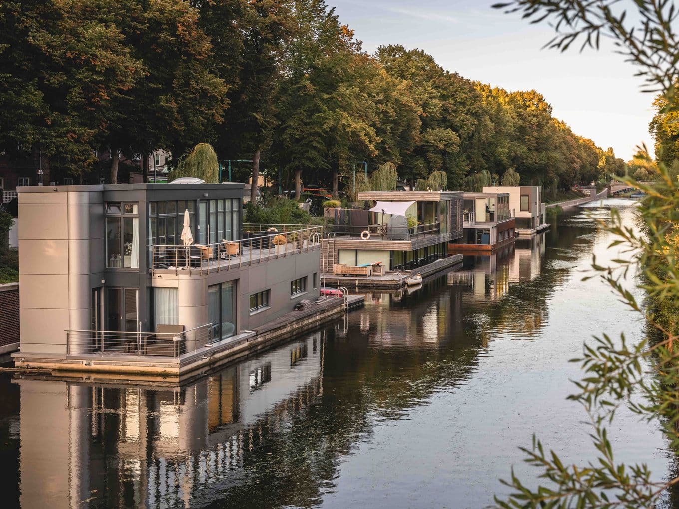 Modern houseboats in Wandsbek, lined up along a quiet canal, surrounded by trees and greenery under a clear sky.