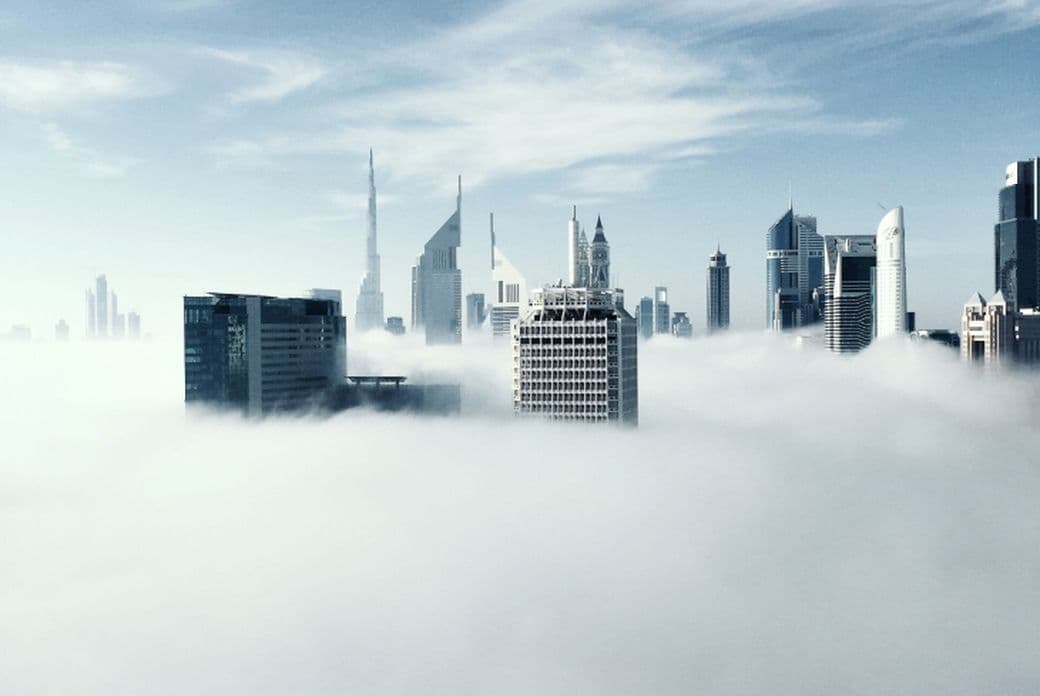 Skyscrapers emerging through thick fog under a blue sky, creating a surreal cityscape.