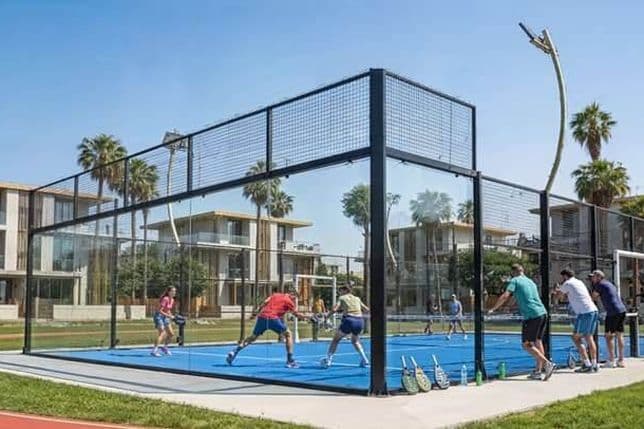 People playing paddle tennis on a blue court surrounded by glass walls, with spectators watching, set against a backdrop of palm trees and modern buildings.