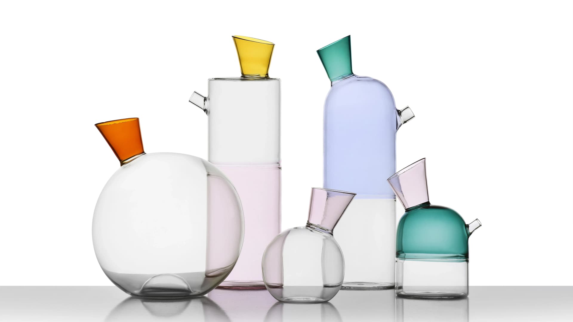 Colorful, geometrically shaped glass vases with vibrant spouts in amber, yellow, teal, and pink, arranged against a white background.