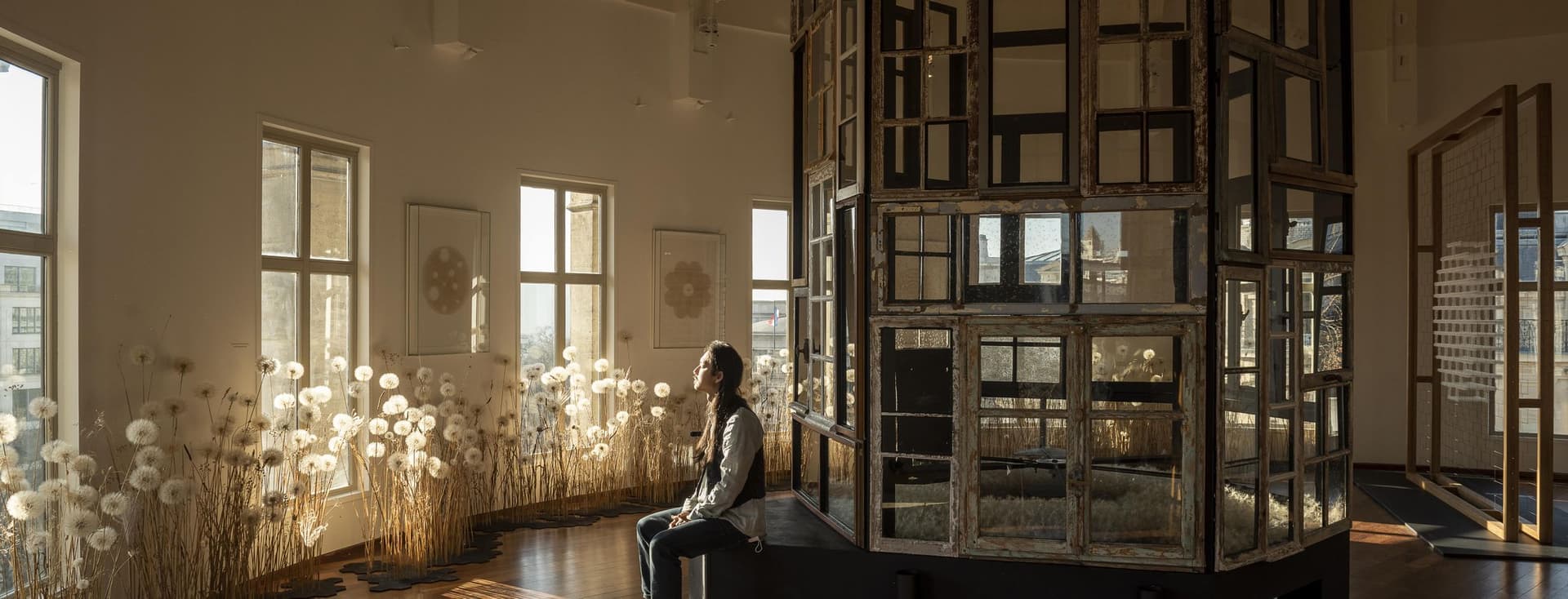 A woman sits in a sunlit room with window art. The room has wood floors and white walls.