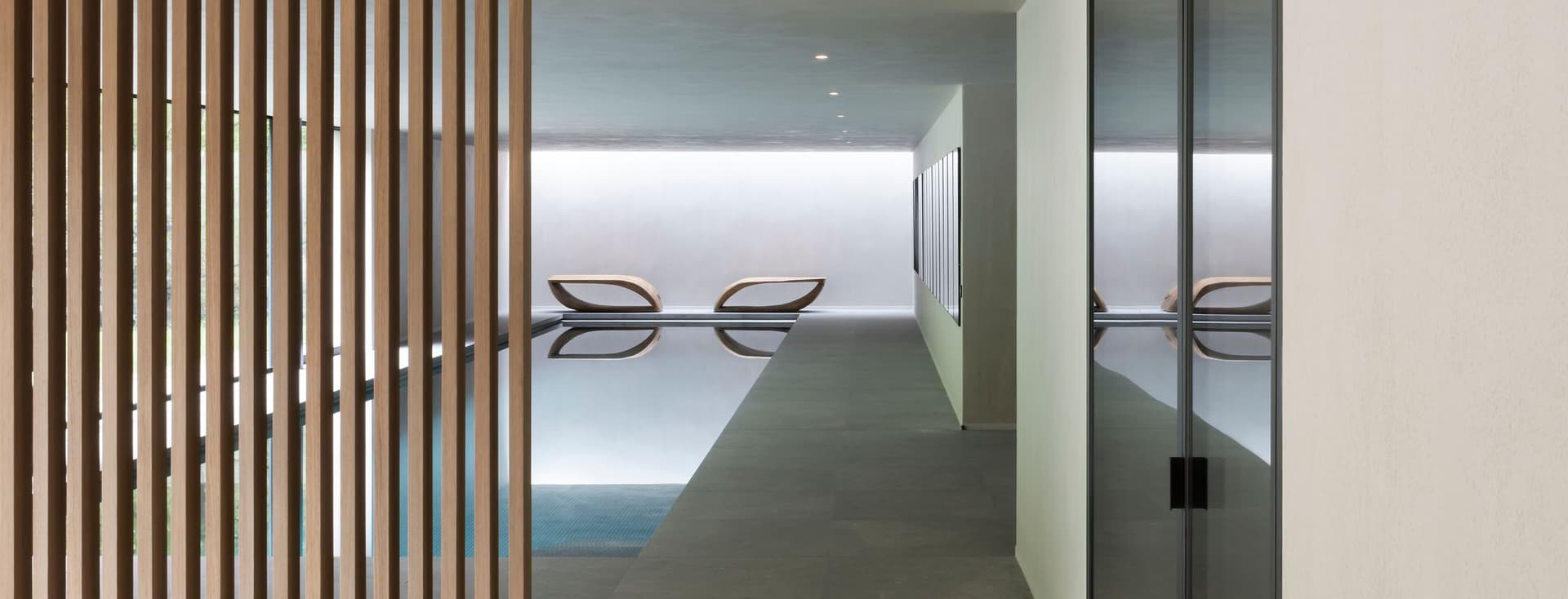 Minimalist indoor pool seen through a corridor; vertical wooden slats on the left, glass wall on the right, and two lounge chairs by the pool.