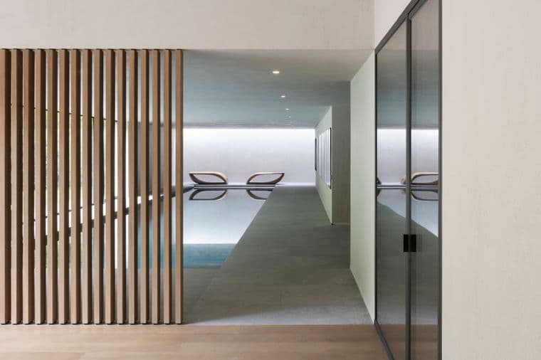 Minimalist indoor pool seen through a corridor; vertical wooden slats on the left, glass wall on the right, and two lounge chairs by the pool.