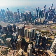 Aerial view of Dubai with numerous skyscrapers, highways, and a distant coastline under a clear sky.