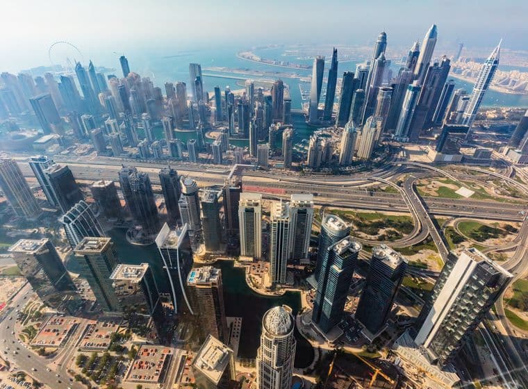 Aerial view of Dubai with numerous skyscrapers, highways, and a distant coastline under a clear sky.