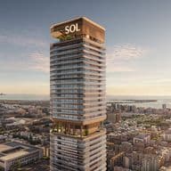 Tall modern skyscraper labeled "SOL" with terraces, overlooking a cityscape and ocean, with a famous sail-shaped hotel in the background.