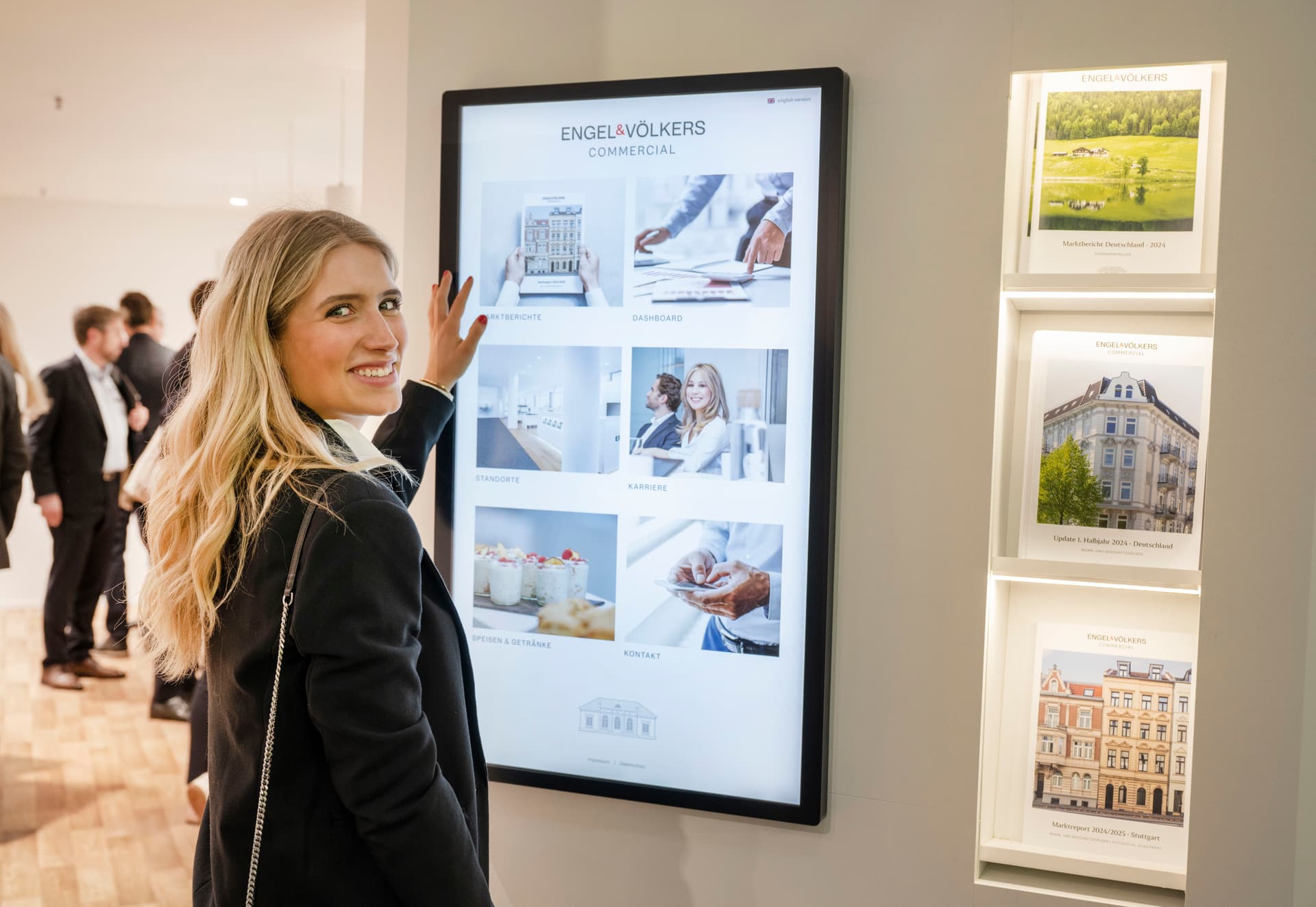 Woman interacting with a touchscreen display in a modern office, showcasing real estate images. Others are visible in the background.