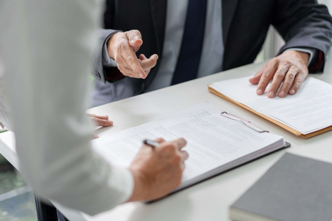 Two people in formal attire discussing documents at a table, one writing on a clipboard, the other gesturing with a hand.