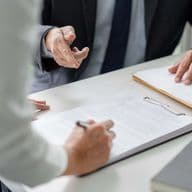 Two people in formal attire discussing documents at a table, one writing on a clipboard, the other gesturing with a hand.