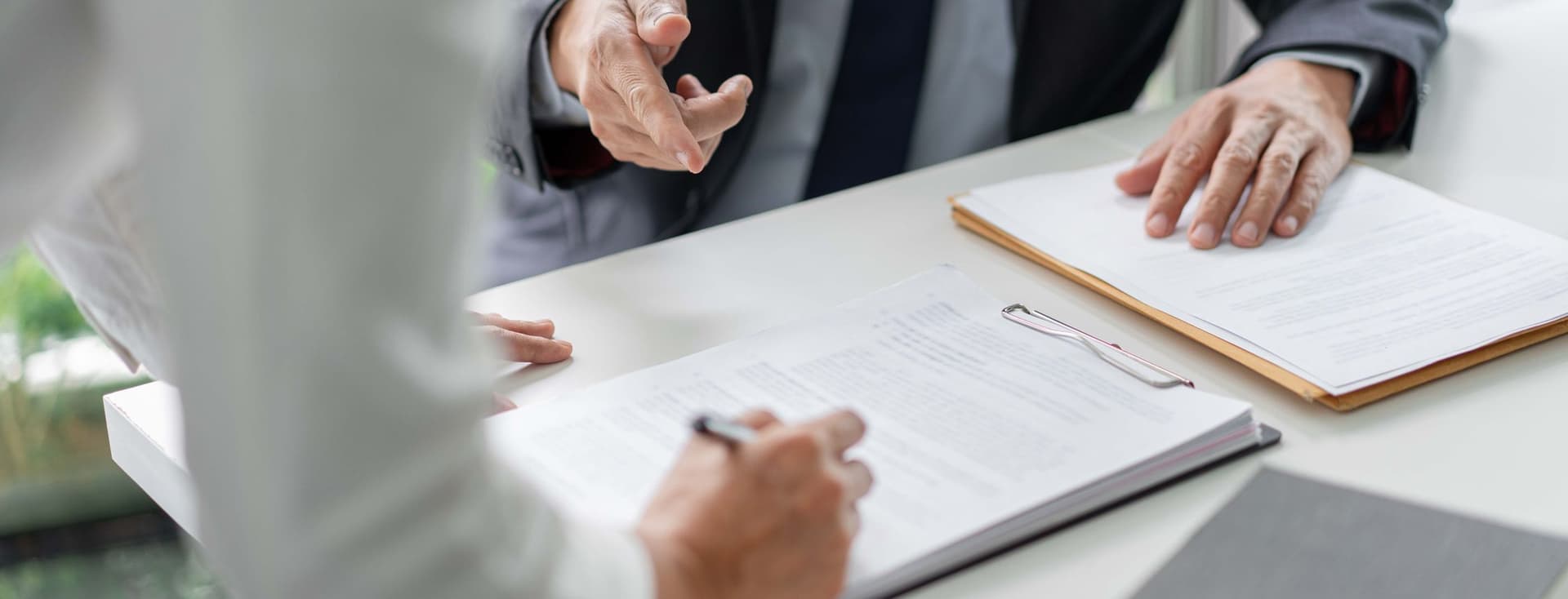 Two people in formal attire discussing documents at a table, one writing on a clipboard, the other gesturing with a hand.