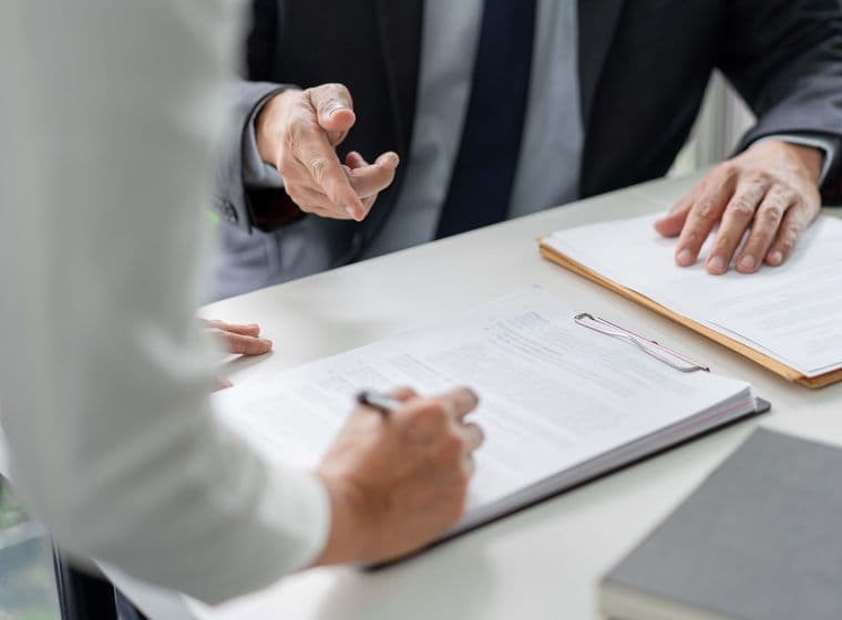 Two people in formal attire discussing documents at a table, one writing on a clipboard, the other gesturing with a hand.