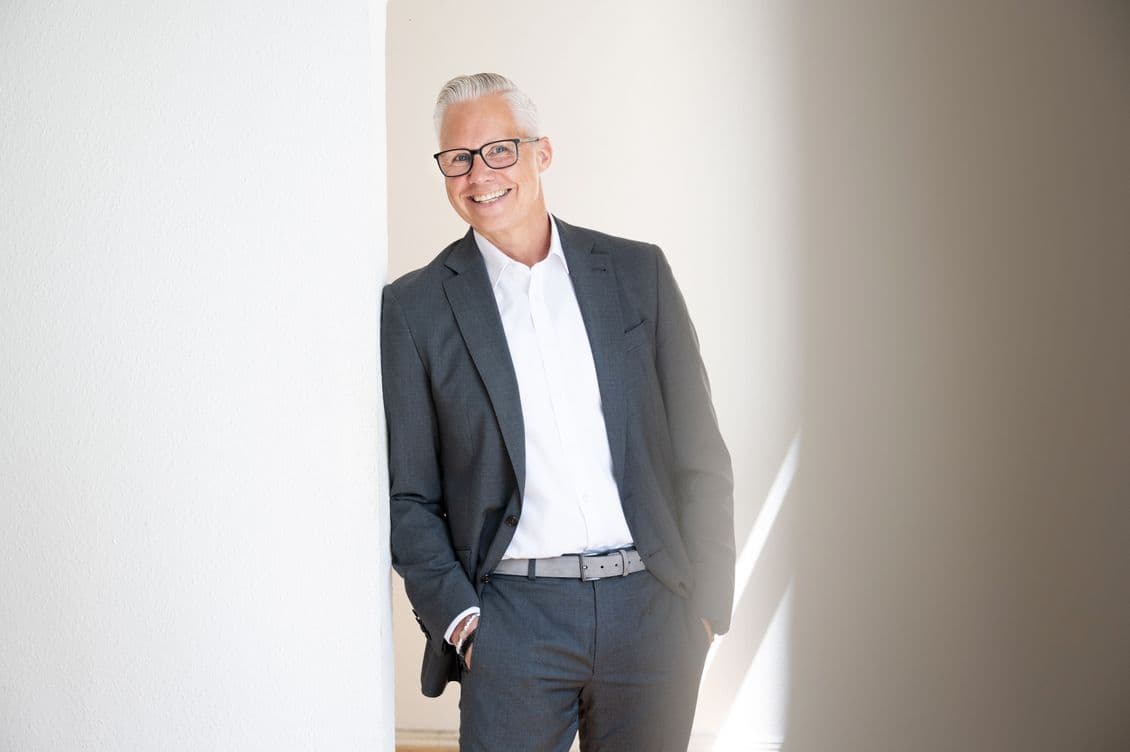 beaming best ager in a gray suit leaning against a white wall