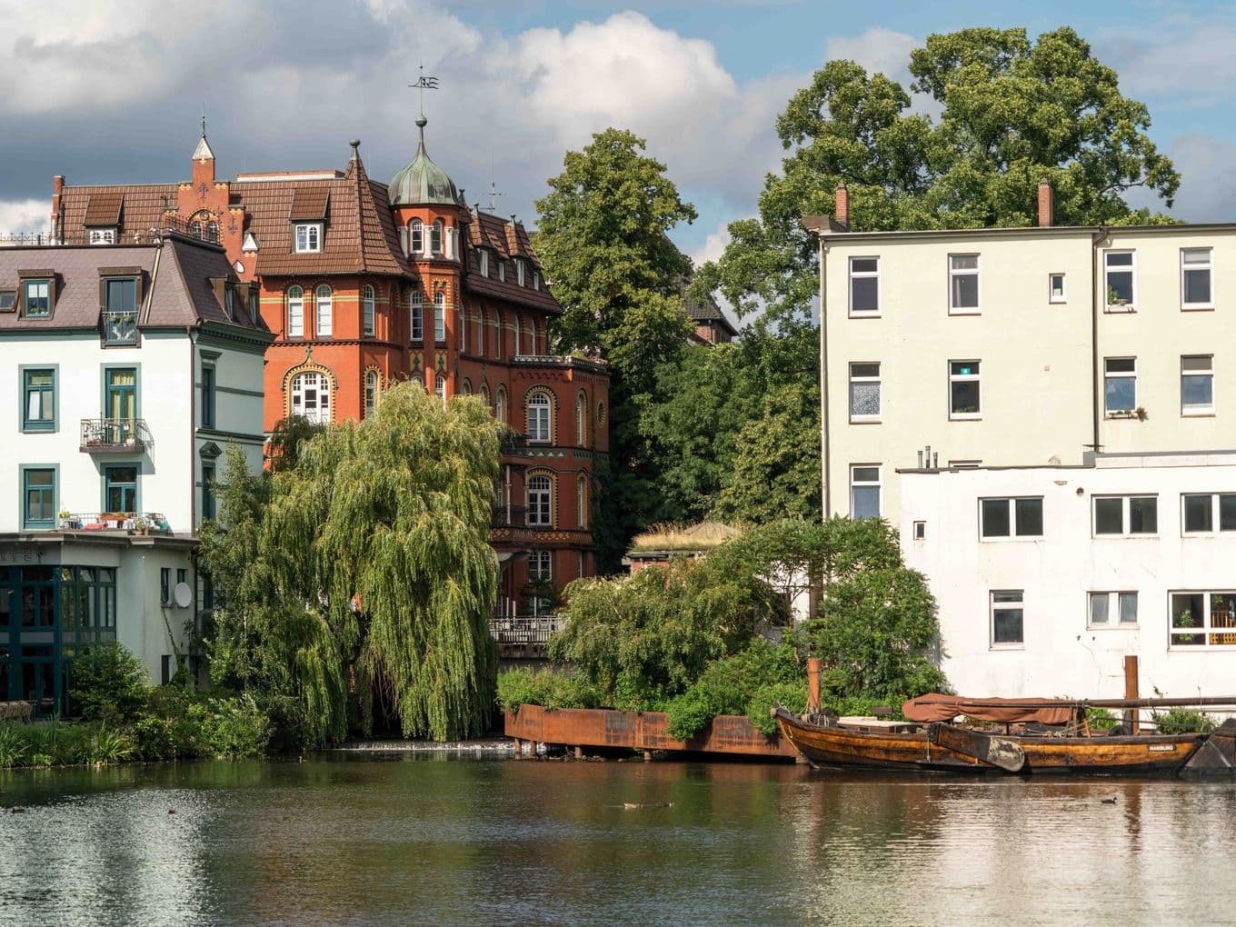 Historic buildings on a quiet river in Bergedorf, with lush greenery and a small docked boat under a partly cloudy sky.