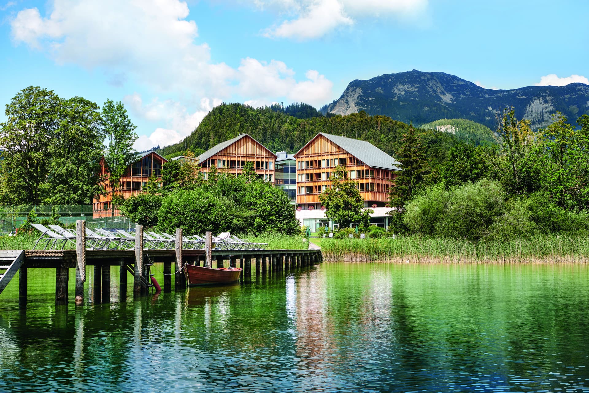 Maisons en bois aux toits gris nichées dans des arbres verts, avec des montagnes et un ciel bleu avec des nuages en arrière-plan.