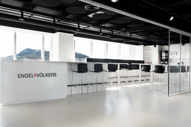 Εσωτερικό γραφείου της Engel & Völkers με έναν λευκό πάγκο, μαύρα σκαμπό και θέα σε ένα βουνό μέσα από μεγάλα παράθυρα.