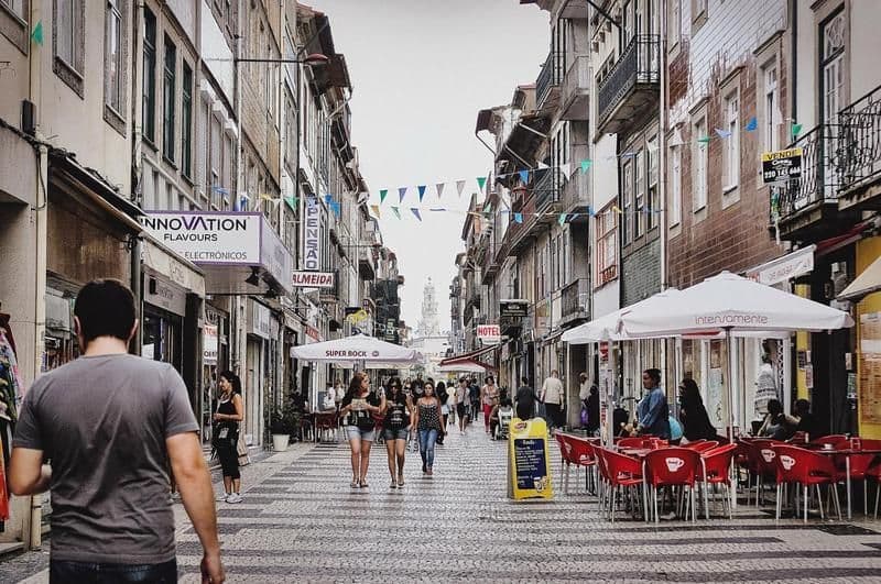 A bustling pedestrian street with people walking, outdoor cafes, colorful banners overhead, and shops lining both sides.