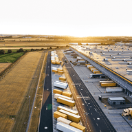Luftaufnahme eines großen Logistikzentrums mit zahlreichen Lkw und Anhängern auf geordneten Ladebereichen, angrenzend an weite Felder bei tiefstehender Sonne.