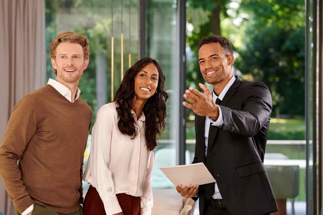 Three people smiling, standing indoors near large windows. One person in a suit gestures while holding documents. Lush greenery visible outside.