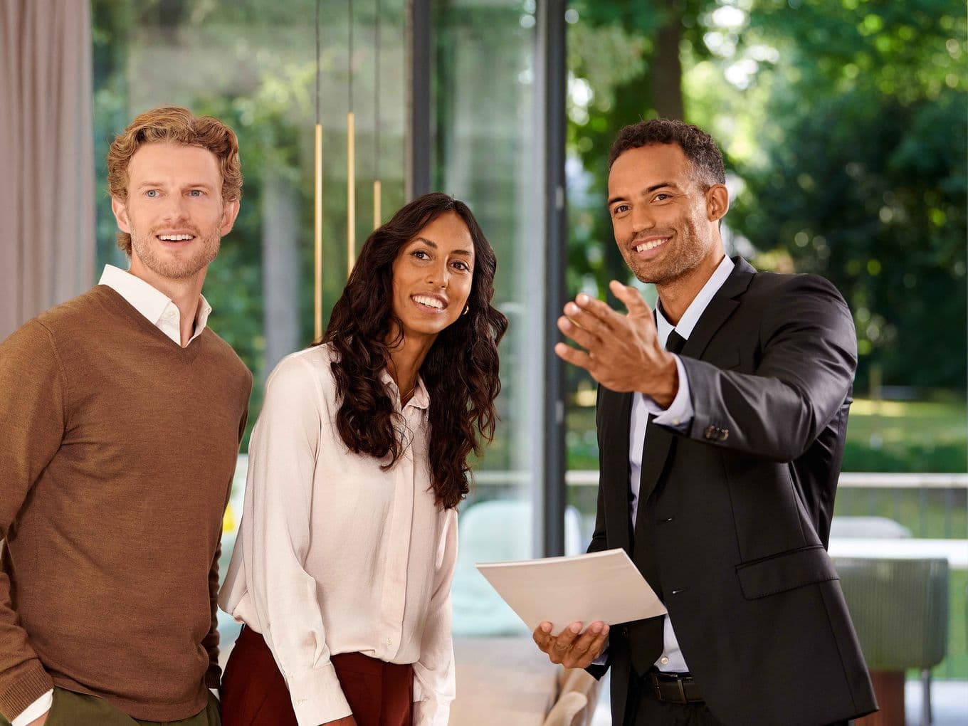 Three people smiling, standing indoors near large windows. One person in a suit gestures while holding documents. Lush greenery visible outside.