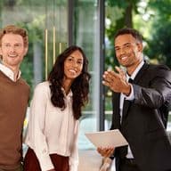 Three people smiling, standing indoors near large windows. One person in a suit gestures while holding documents. Lush greenery visible outside.