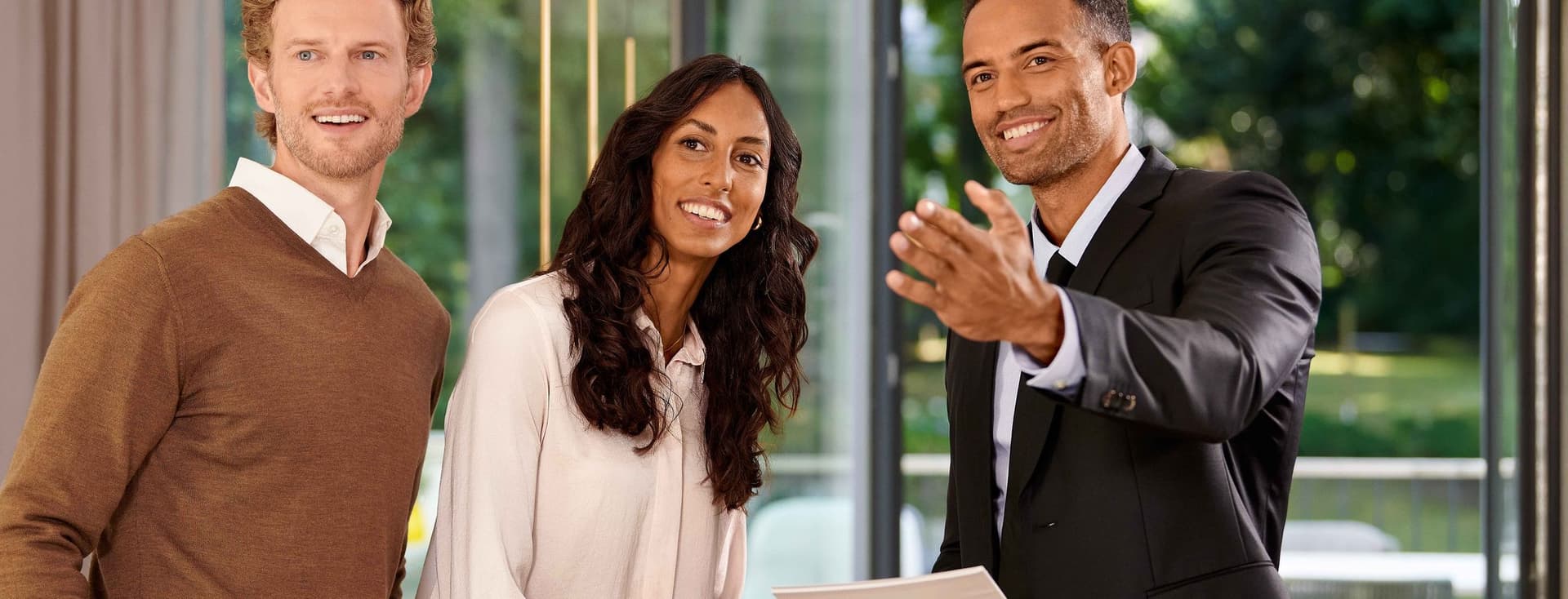 Three people smiling, standing indoors near large windows. One person in a suit gestures while holding documents. Lush greenery visible outside.