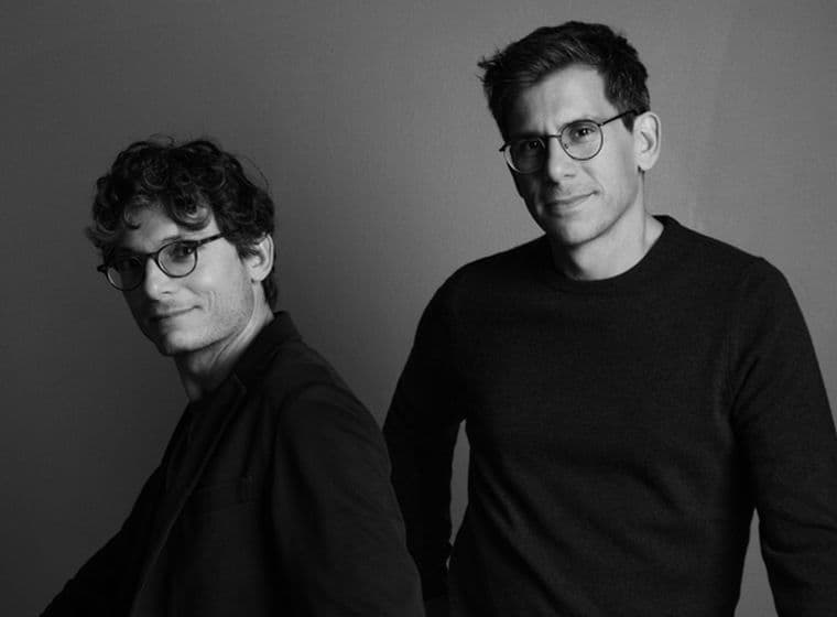 Μαύρο και άσπρο πορτρέτο δύο ανδρών που φορούν γυαλιά και σκούρα ρούχα σε γκρι φόντο.