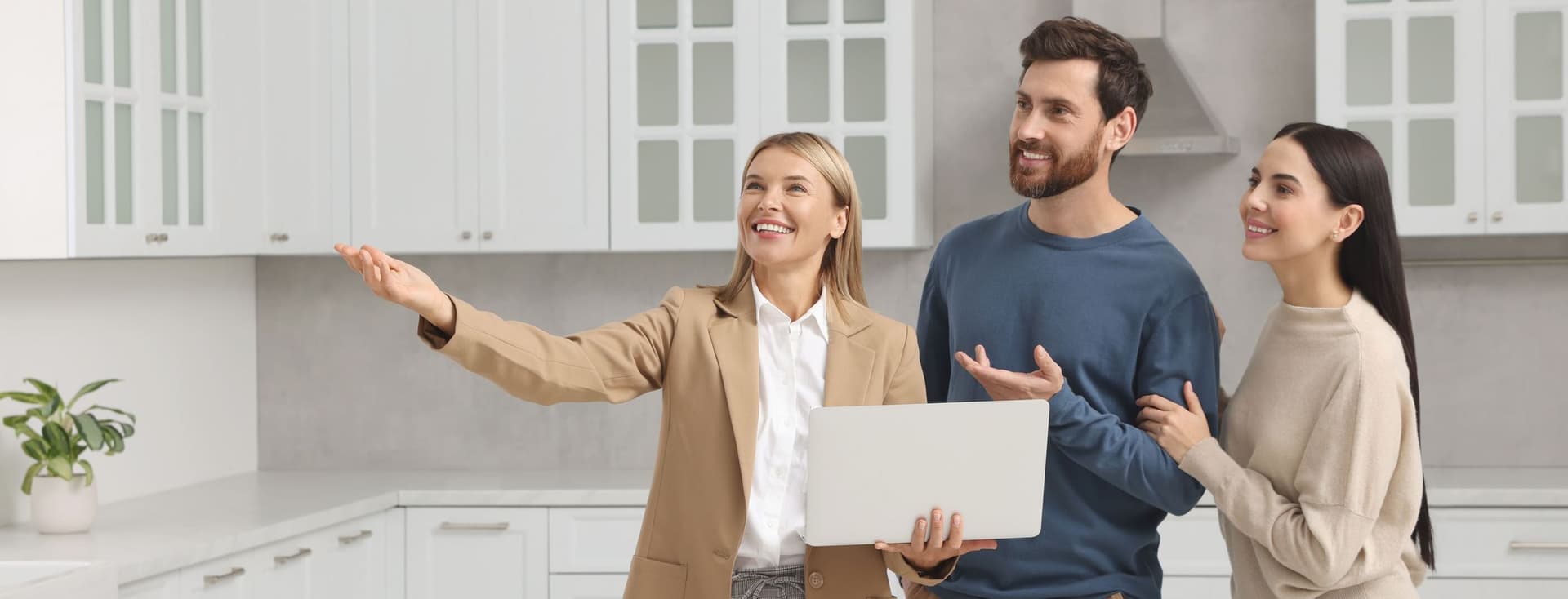 A real estate agent shows a couple a modern white kitchen, holding a laptop and gesturing to the space.
