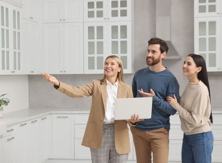 A real estate agent shows a couple a modern white kitchen, holding a laptop and gesturing to the space.