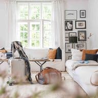 Cozy living room with white sofas, assorted pillows, a suitcase, and a travel bag. Large window and framed art on walls.