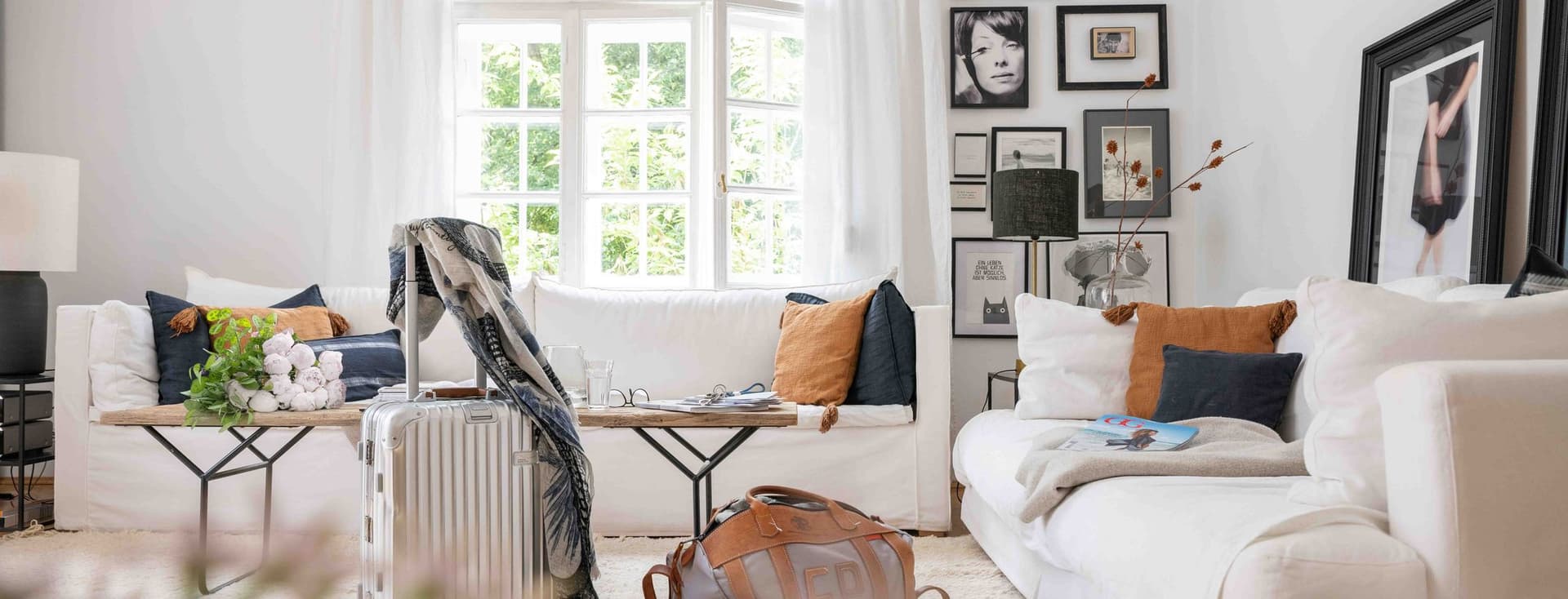 Cozy living room with white sofas, assorted pillows, a suitcase, and a travel bag. Large window and framed art on walls.
